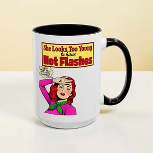 May include: White ceramic mug with a black interior and handle. The mug features a comic-style illustration of a woman with red hair and the text "She Looks Too Young to have Hot Flashes."