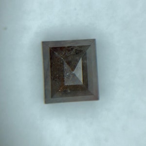 May include: A square-cut, dark gray gemstone with a faceted surface. The gem has a central, square-shaped facet and a slightly textured appearance. The stone is set against a white background.