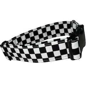 May include: A pet collar featuring a black and white checkered pattern. The collar is constructed from fabric and includes a black plastic side-release buckle, a black plastic adjustment slider, and a metal D-ring for attaching a leash or tags.