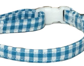 May include: A blue and white gingham patterned collar featuring a white plastic side-release buckle, a white plastic adjustable slider, and a small silver-toned metal bell attached to a split ring. The collar is constructed from fabric.
