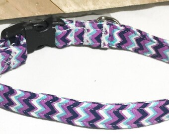 May include: A pet collar featuring a repeating chevron pattern in shades of purple, dark blue, light blue, and white. The collar includes a black plastic side-release buckle, a black plastic sliding adjuster, and a silver-toned metal D-ring for a leash.