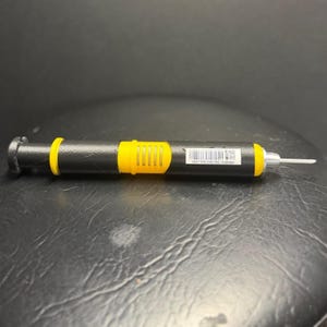 Puede incluir: Un destornillador de precisión negro y amarillo con punta plateada. El destornillador tiene un mango negro con una sección de agarre amarilla y un anillo amarillo cerca de la punta. Un código de barras y números están impresos en el mango.