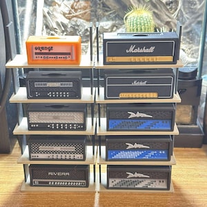 May include: A collection of miniature guitar amplifiers displayed on a multi-tiered shelf. The amps feature various designs, including the "Marshall" and "Orange" brands, with black, orange, and silver color schemes. The shelf is made of light-colored wood.