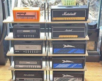 May include: A collection of miniature guitar amplifiers displayed on a multi-tiered shelf. The amps feature various designs, including the "Marshall" and "Orange" brands, with black, orange, and silver color schemes. The shelf is made of light-colored wood.