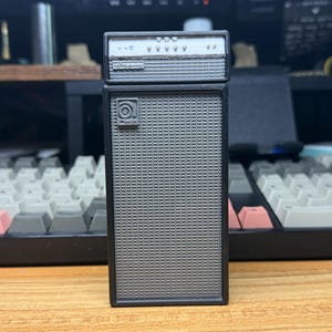 Puede incluir: Un amplificador de guitarra en miniatura de color gris que consta de una caja de altavoz rectangular con una rejilla frontal texturizada y una unidad de cabezal más pequeña encima. El cabezal muestra varios botones y un logo.