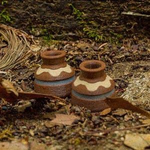 Peut inclure: Deux petits pots en argile brune texturée avec une large ouverture circulaire au sommet. Chaque pot présente une bande horizontale décorative de motifs festonnés couleur crème près du bord et une rayure bleu clair sur le corps inférieur.