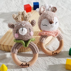 May include: Two handmade crochet baby rattles. One is a deer with antlers, the other a deer with closed eyes. Both have wooden rings with names. The deer rattles are brown, white, pink, and green. Colorful wooden blocks are scattered around the rattles.