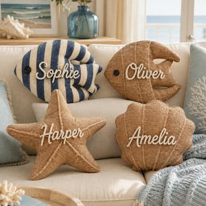 Könnte beinhalten: Vier dekorative Kissen in Meeresbewohner-Form. Ein blau-weiß gestreifter Fisch mit der Aufschrift Sophie, ein beiger Fisch mit der Aufschrift Oliver, ein beiger Seestern mit der Aufschrift Harper und eine beige Muschel mit der Aufschrift Amelia.