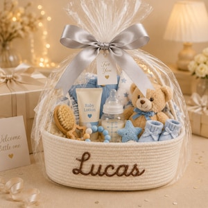 Könnte beinhalten: Ein weißer geflochtener Korb mit dem in Braun gestickten Namen Lucas, der ein Baby-Geschenkset enthält. Zu den Artikeln gehören ein beiger Teddybär, blaue Babyschuhe, eine Baby-Lotion, ein Fläschchen, eine Holzbürste und ein sternförmiges Spielzeug, verpackt in transparenter Folie mit einem silbernen Band und einem Anhänger mit der Aufschrift For You.