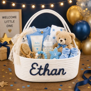 May include: A white rope basket with the name Ethan embroidered in navy blue script. The basket contains a blue ribbed shirt, a baby lotion tube, a baby bottle, a tan teddy bear with a blue bow, blue booties, a wooden brush, and a star toy.