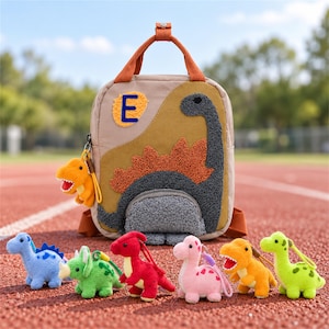Könnte beinhalten: Ein beigefarbener Rucksack mit einer texturierten Dinosaurier-Applikation und einem blauen Buchstaben E als Aufnäher, dazu sechs kleine Plüsch-Dinosaurier-Schlüsselanhänger in Blau, Grün, Rot, Rosa, Orange und Limettengrün mit Schlaufe.