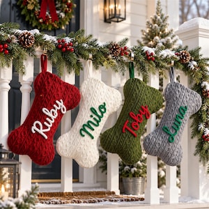 May include: Four bone-shaped Christmas stockings made of cable-knit fabric in red, white, olive green, and gray. Each stocking features a name embroidered in a contrasting color: Ruby in white, Milo in green, Toby in red, and Luna in green.