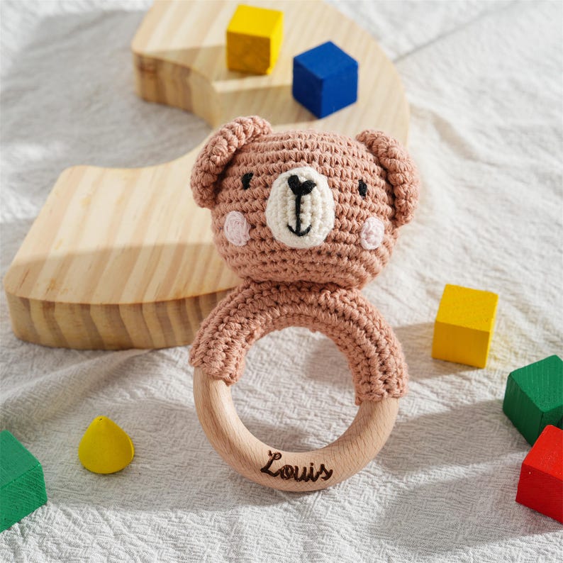 May include: A handmade, crochet bear-shaped baby rattle in light brown with a wooden ring. The rattle features a white muzzle, black eyes, and the name "Louis" engraved on the wooden ring. Colorful wooden blocks are scattered around the rattle.