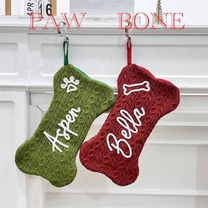 May include: Two bone-shaped Christmas stockings made of cable-knit fabric. The left stocking is green with a white paw print and the name Aspen. The right stocking is red with a white bone icon and the name Bella. Both have hanging loops.