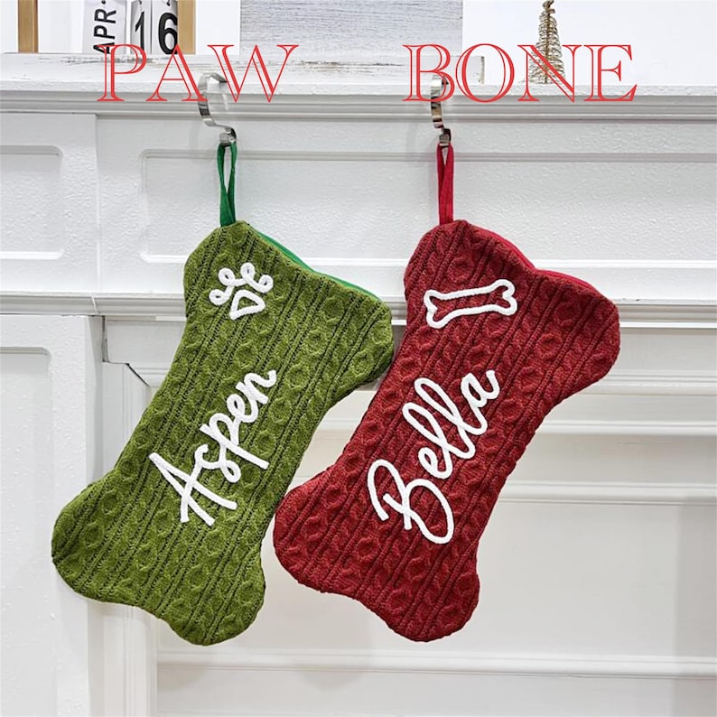 May include: Two bone-shaped Christmas stockings made of cable-knit fabric. The left stocking is green with a white paw print and the name Aspen. The right stocking is red with a white bone icon and the name Bella. Both have hanging loops.