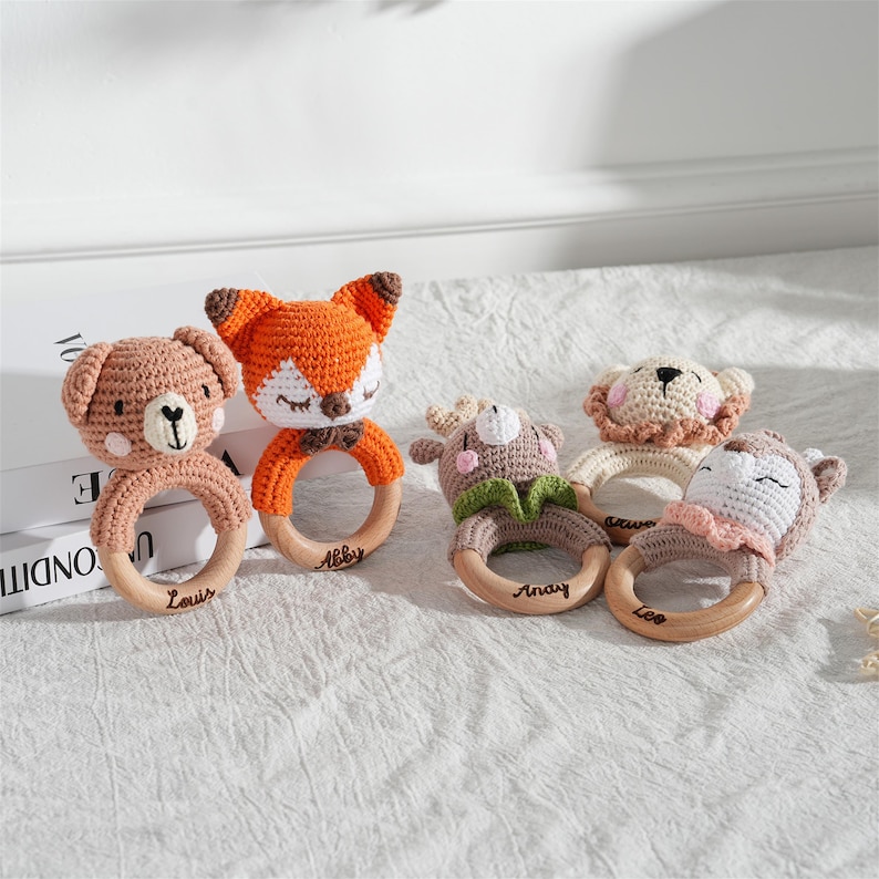 May include: A collection of five handmade crochet baby rattles. The rattles feature animal designs, including a bear, fox, deer, lion, and sheep, with wooden ring handles. Each rattle is a different color and has a name on the wooden ring.