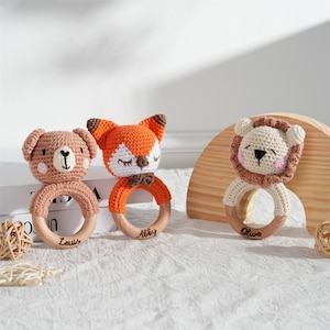 May include: Three crochet baby rattles in the shape of a bear, fox, and lion. Each rattle has a wooden ring base and is personalized with a name. The bear is brown, the fox is orange and white, and the lion is beige.