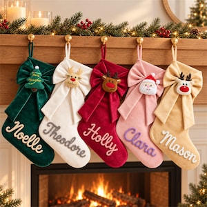 May include: Five velvet Christmas stockings in dark green, white, red, light pink, and tan. Each stocking features a large fabric bow, a plush holiday character or tree ornament, and a name embroidered in cursive script: Noelle, Theodore, Holly, Clara, and Mason.