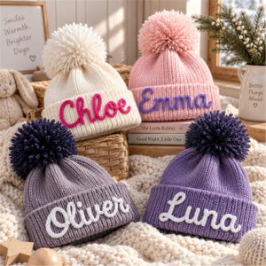 Puede incluir: Cuatro gorros de punto con pompones, cada uno con un nombre personalizado en letra cursiva. El gorro crema muestra el nombre Chloe en rosa, el gorro rosa muestra Emma en morado, el gorro gris presenta a Oliver en blanco y el morado dice Luna en blanco.