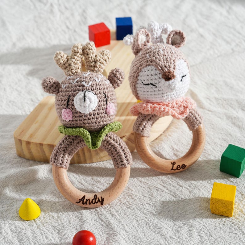 May include: Two handmade crochet baby rattles. One is a deer with antlers, the other a deer with closed eyes. Both have wooden rings with names. The deer rattles are brown, white, pink, and green. Colorful wooden blocks are scattered around the rattles.