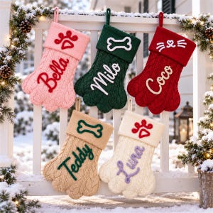 May include: Five paw-shaped knit Christmas stockings in pink, green, red, tan, and cream. Each stocking features a cuff with an embroidered icon and a name: Bella with a paw print, Milo with a bone, Coco with whiskers, Teddy with a bone, and Luna with a paw.