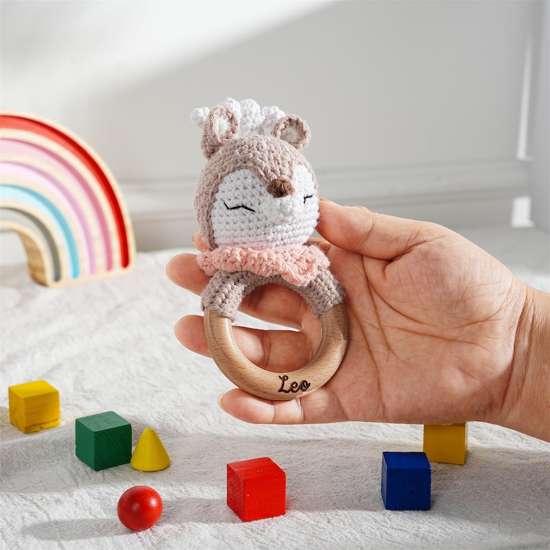 May include: A handmade crochet baby rattle in the shape of a deer, held in a hand. The rattle features a wooden ring and the name "Leo" engraved on it. The deer is light brown, white, and pink. Colorful wooden blocks are scattered around.