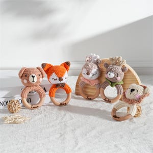 May include: A collection of five handmade crochet baby rattles. Each rattle features a different animal head: a bear, fox, deer, deer with antlers, and a lion. The animal heads are attached to wooden rings. The names "Louis", "Abby", "Lee", "Andy", and "Olive" are engraved on the rings.