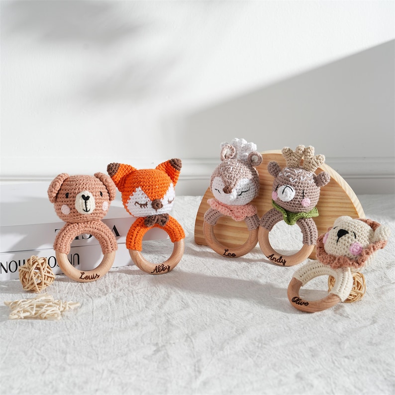 May include: A collection of five handmade crochet baby rattles. Each rattle features a different animal head: a bear, fox, deer, deer with antlers, and a lion. The animal heads are attached to wooden rings. The names "Louis", "Abby", "Lee", "Andy", and "Olive" are engraved on the rings.