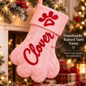 May include: A light pink knit Christmas stocking shaped like a paw, featuring a red yarn paw print on the cuff and the name Clover in red raised yarn script. The text on the image reads Handmade Raised Yarn Name and Soft 3D yarn detail.