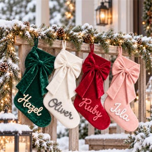 May include: Four velvet Christmas stockings hanging in a row, each featuring a large decorative bow at the top and a name embroidered in script. The stockings are colored dark green, cream, deep red, and light pink, with names Hazel, Oliver, Ruby, and Isla.