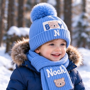 Op de afbeelding: Een lichtblauwe gebreide muts met een bijpassende pompon en een rechthoekige patch met een berengezicht en de letters B, Y en N. Een bijpassende lichtblauwe gebreide sjaal is voorzien van de naam Noah in witte letters en een berengezicht-patch.