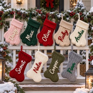 May include: A collection of nine cable-knit Christmas stockings hanging on a white railing. Five are paw-shaped with names Daisy, Max, Olive, Ruby, and Archie. Four are bone-shaped with names Scout, Nala, Cooper, and Finn in various colors.