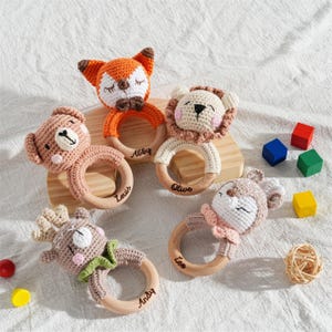 May include: A collection of five handmade crochet baby rattles in the shape of animals, including a fox, lion, bear, deer, and another deer. Each rattle has a wooden ring and is personalized with a name. Colorful wooden blocks are also in the image.