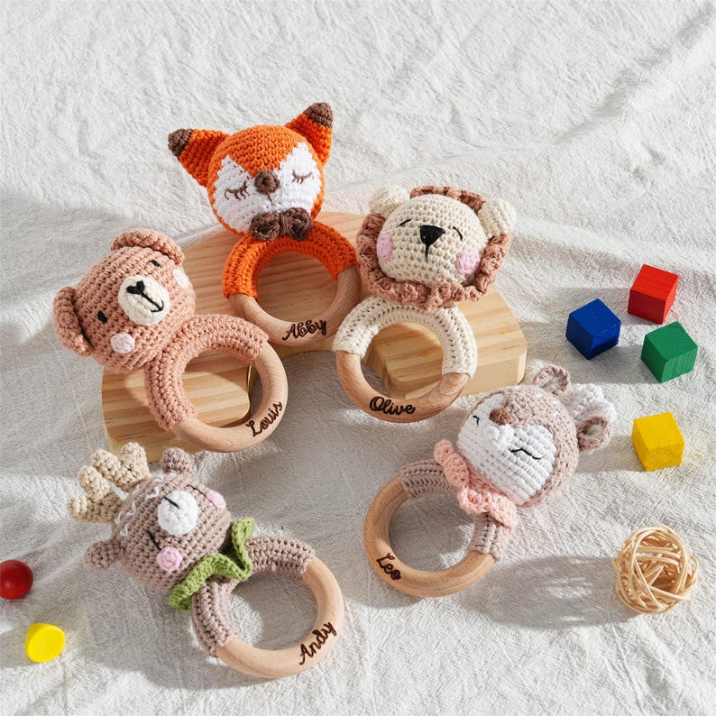 May include: A collection of five handmade crochet baby rattles in the shape of animals, including a fox, lion, bear, deer, and another deer. Each rattle has a wooden ring and is personalized with a name. Colorful wooden blocks are also in the image.