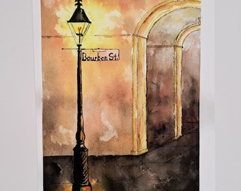 Puede incluir: Pintura a la acuarela de Bourbon Street al anochecer. Una farola negra con un letrero que dice "Bourbon St." se ilumina contra un cielo cálido y anaranjado. Arcos y una calle adoquinada completan la escena.