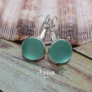 May include: A pair of silver dangle earrings with round, frosted aqua blue glass cabochons. The earrings are on a wooden surface with the word "Aqua" written below them.
