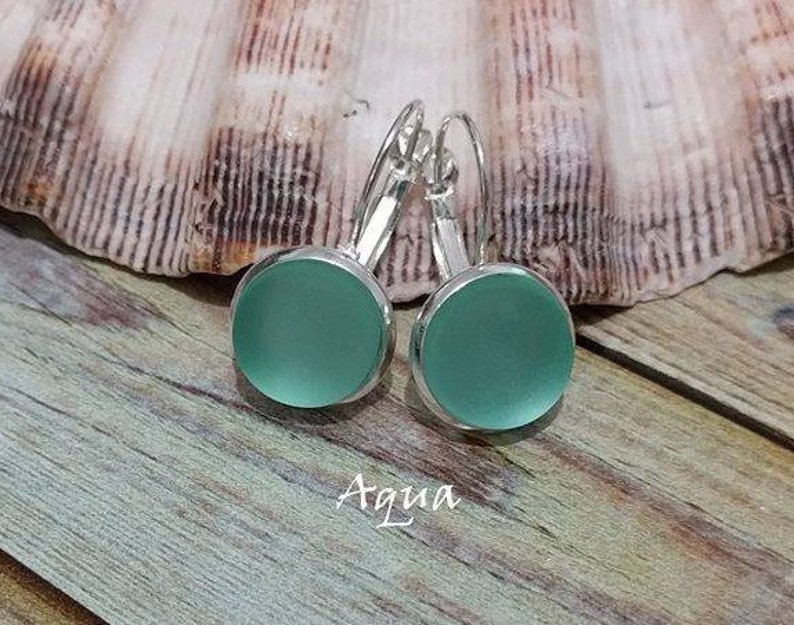 May include: A pair of silver dangle earrings with round, frosted aqua blue glass cabochons. The earrings are on a wooden surface with the word "Aqua" written below them.