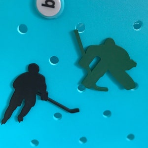 May include: Two black silhouettes of hockey players on a bright blue background. One player is holding a hockey stick and the other is in a goalie stance.