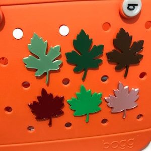 May include: Six colorful leaf-shaped charms in various shades of green, red, brown, and pink. The charms are arranged on an orange background with a small white circle in the upper right corner.