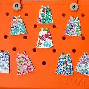 May include: A bright orange tote bag with the word "BOGG" printed on the bottom. The bag has eight small, colorful, patterned dresses attached to it. The dresses have various designs, including floral, nautical, and abstract patterns.