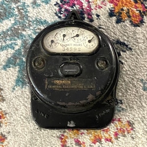 May include: A vintage black General Electric kilowatt-hour meter featuring a circular dial display with multiple small clock-like gauges, the text KILOWATT HOURS 34892, and the words LINE and LOAD embossed on the lower section of the metal casing.