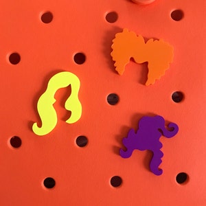 May include: Three felt hair pieces in different colors and shapes. The hair pieces are yellow, orange, and purple. The hair pieces are on a bright orange background.