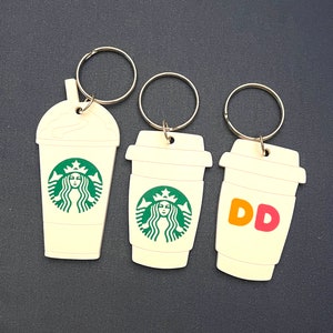 May include: Three cream-colored keychains shaped like coffee cups. One keychain features the Starbucks logo, another the Starbucks logo, and the third has the letters "DD" in orange and pink. Each keychain has a silver ring.