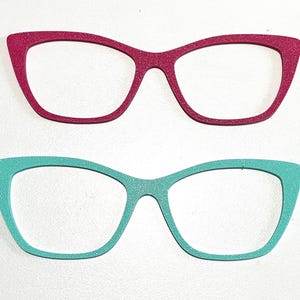 May include: Two pairs of glittery eyeglasses frames. One pair is a deep pink color, and the other is a bright teal. Both pairs have a cat-eye shape. The frames are set against a white background.