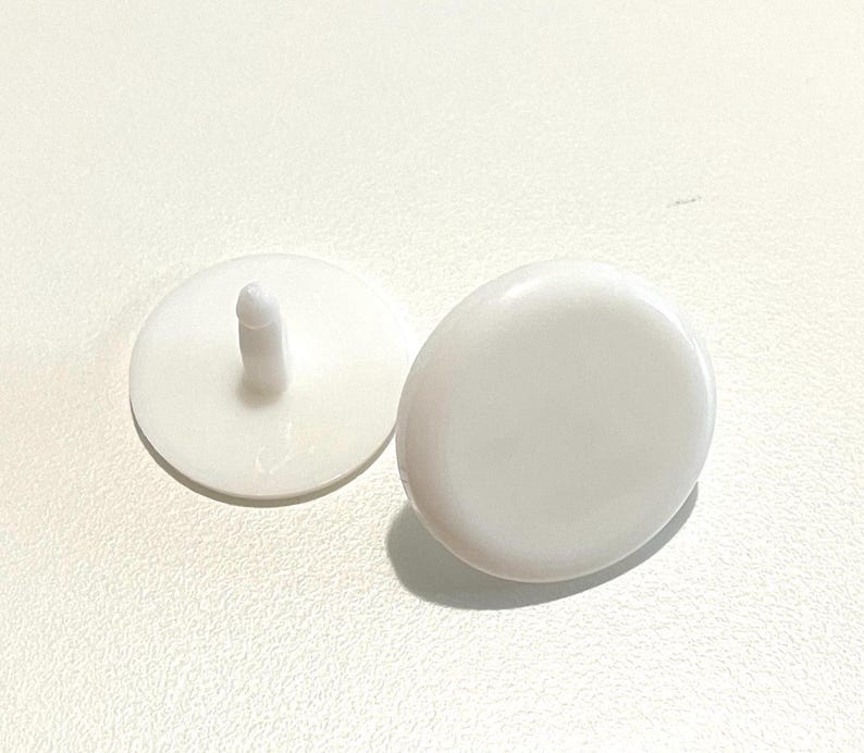 May include: Two white plastic buttons with a small raised nub on one button. The buttons are round and smooth.