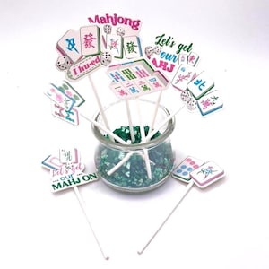 May include: A collection of Mahjong-themed party decorations. The toppers feature colorful tiles, dice, and phrases like "Mahjong" and "Let's get our AHJ." They are displayed in a clear glass jar filled with green decorative stones, perfect for a game night.