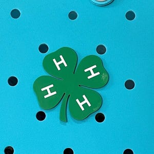 May include: A green four-leaf clover charm with white "H" letters on each leaf. The charm is set against a bright blue background with evenly spaced black circular cutouts. The clover is a symbol of good luck.