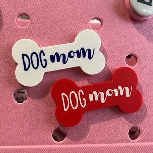 May include: Two bone-shaped charms with the text "DOG mom" on them. One charm is white with blue text and the other is red with white text.