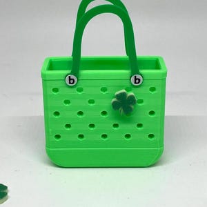 May include: A small green tote bag with a clover charm. The bag has a white "b" on each handle.