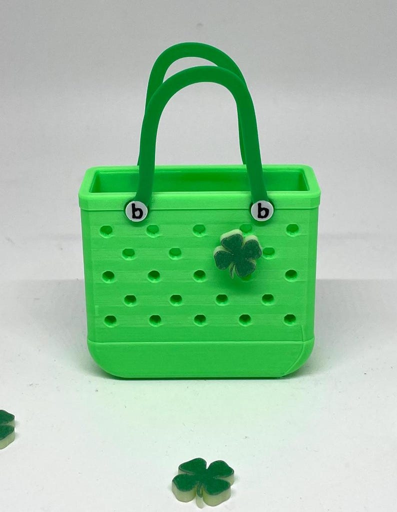 May include: A small green tote bag with a clover charm. The bag has a white "b" on each handle.